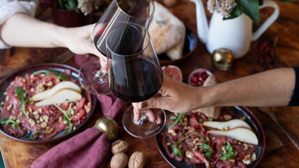 Zwei Personen stoßen mit Rotwein an. Auf dem Tisch befinden sich zwei Teller mit Essen.