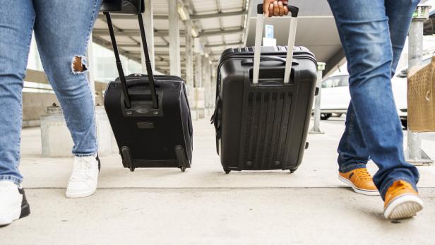 Zwei Personen mit Rollkoffern stehen auf einem Bahnsteig oder Flughafen.