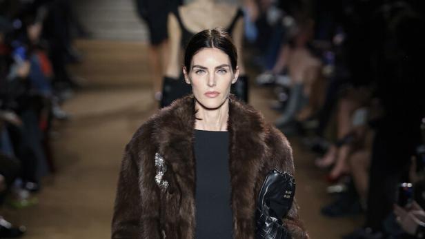 Eine Frau in einem braunen Pelzmantel und schwarzem Kleid läuft über einen Laufsteg.