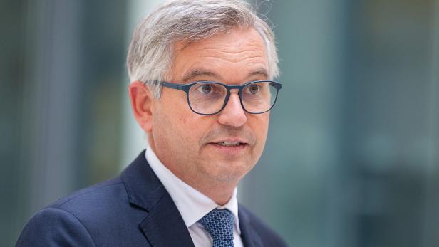 Ein Mann mit grauen Haaren, Brille und Anzug spricht, unscharfer Hintergrund.