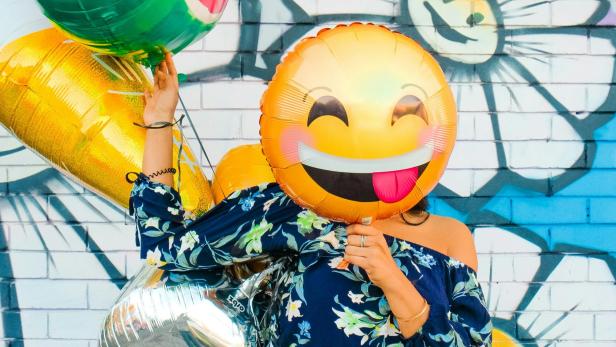 Eine Frau, die sich einen Ballon mit Emoji-Motiv vor das Gesicht hält.