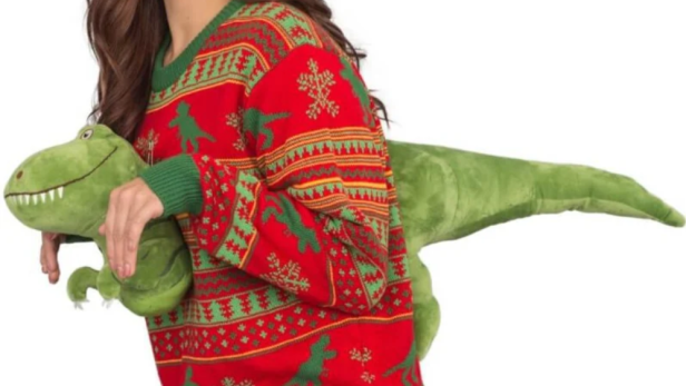 Eine Frau trägt einen roten Weihnachtspullover mit Dinosaurier-Motiv und hält ein grünes Dinosaurier-Stofftier.