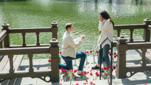 Mann macht Frau kniend vor einer Brücke einen Heiratsantrag