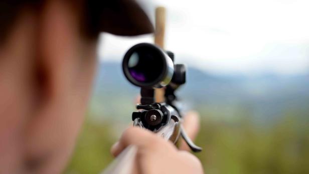 Eine Person zielt mit einem Gewehr samt Zielfernrohr in eine unscharfe Landschaft.