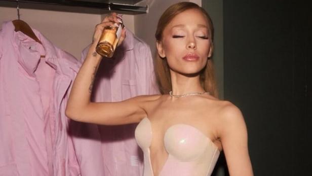 Sängerin Ariana Grande hat ein Parfum in der Hand, mit dem sie sich mit geschlossenen Augen besprüht.