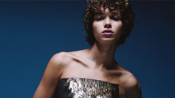 Eine Frau in einem trägerlosen, schwarzen Kleid mit silbernen und goldenen Fransen vor blauem Hintergrund.