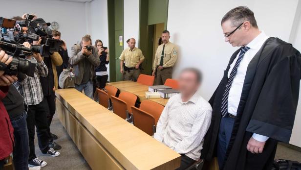 Ein Angeklagter sitzt im Gerichtssaal, während Fotografen und ein Anwalt anwesend sind.