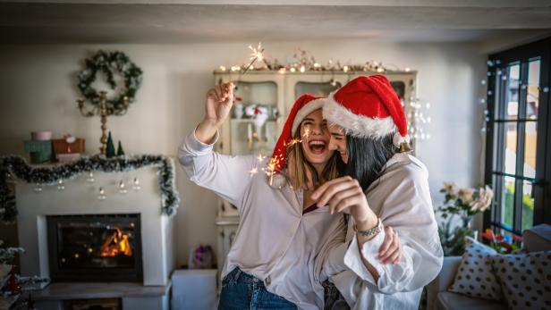 Zwei Frauen mit Weihnachtsmützen feiern mit Wunderkerzen in einem Wohnzimmer.