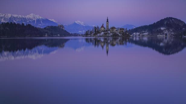 Eine Kircheninsel inmitten des Bleder Sees, aufgenommen am Abend. Rundherum stehen Hügeln und Berge