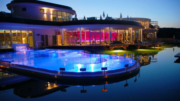 Modernes Wellnessgebäude mit beleuchtetem Außenpool und großen Glasfronten in der Abenddämmerung.