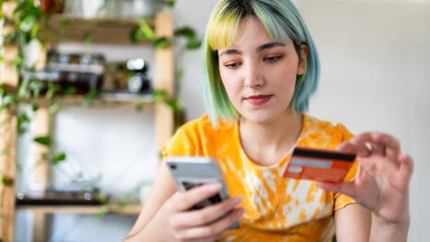 Jugendliche mit kurzen blauen Haaren schaut auf ihr Smartphone und hält eine Kreditkarte in der Hand