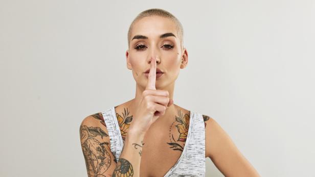 Eine junge Frau mit Tattoos hält den Finger vor den Mund.