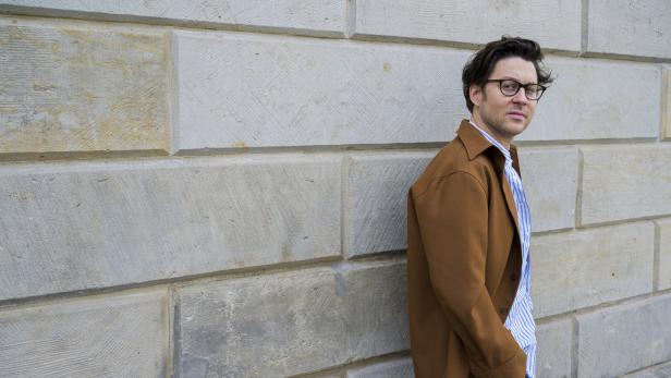 Ein Mann mit Brille und brauner Jacke steht vor einer Steinwand.
