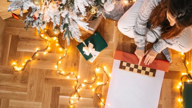 Eine Frau sitzt neben einem Weihnachtsbaum und verpackt ein Schachbrett als Geschenk.