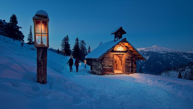Zwei Personen wandern im Schnee zu einer beleuchteten Holzhütte.