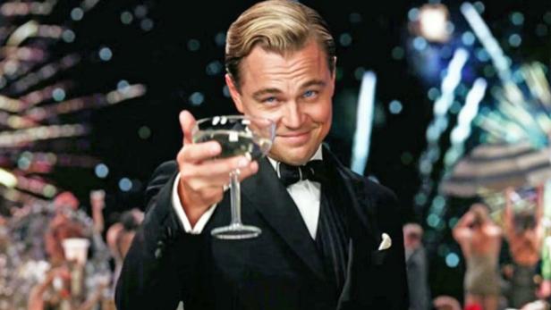 Der große Gatsby im Smoking, gespielt von Leonardo DiCaprio, prostet mit einer Champagnerschale. Im Hintergrund explodiert ein Feuerwerk.