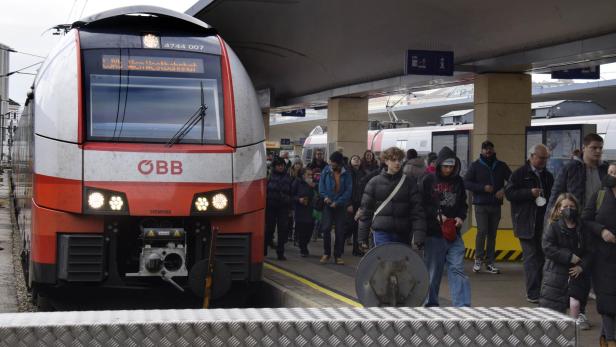 Ein roter Zug steht am Bahnsteig, viele Menschen steigen aus und gehen mit Gepäck und Masken entlang des Bahnsteigs.