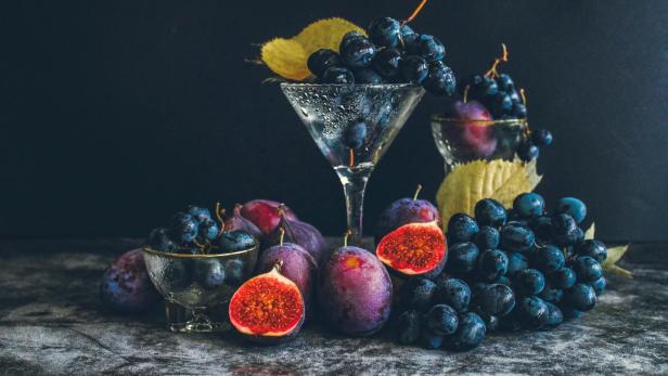 Verschiedene dunkle Trauben, Pflaumen und aufgeschnittene Feigen liegen zusammen mit Blättern auf einer Steinplatte.