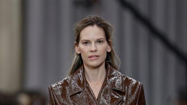Hilary Swank trägt einen glänzenden, braunen Trenchcoat mit Gürtel auf einem Laufsteg.