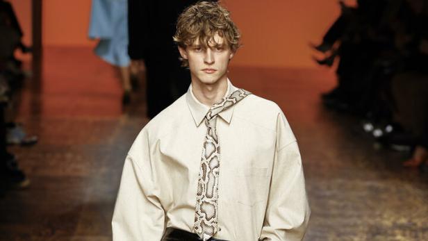 Ein Mann in einem beigen Hemd, einem schwarzen Lackrock und einer grünen Tasche auf einem Laufsteg.