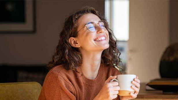 Eine Frau mit lockigen Haaren lächelt der Morgensonne entgegen, während sie eine Tasse Kaffee in der Hand hält. 