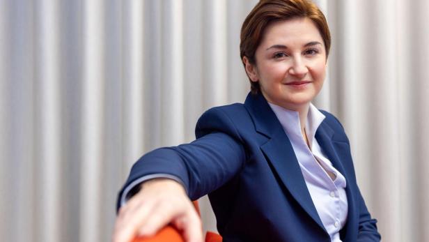 Eine Frau mit kurzem braunem Haar trägt einen blauen Blazer und sitzt entspannt auf einem orangefarbenen Stuhl vor hellen Vorhängen.