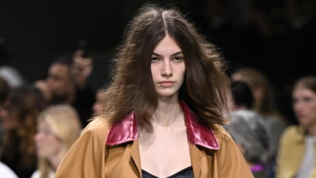 Eine Frau auf dem Laufsteg mit einem Trenchcoat, einem schwarzen Top und einem orangefarbenen Rock.