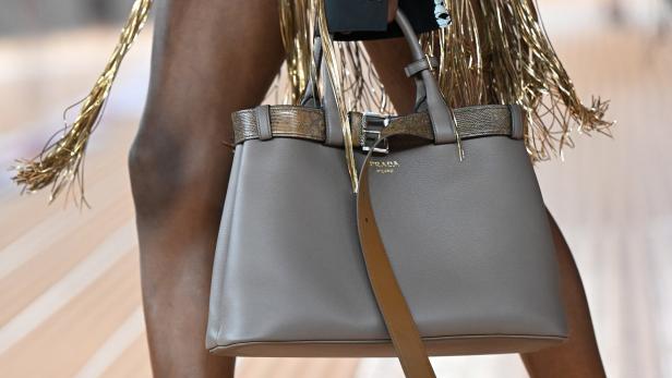 Eine graue Prada-Handtasche mit goldenen Akzenten wird auf einem Laufsteg präsentiert.