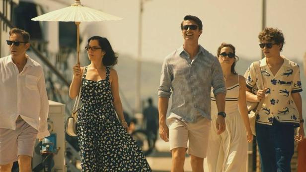 Eine Gruppe von Menschen, möglicherweise aus der Serie „The White Lotus“, geht an einem sonnigen Tag spazieren.