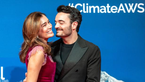 SPECIAL DINNER FOR CLIMATE ACTION/ RED CARPET: ZARRELLA Ein lächelndes Paar steht eng umarmt vor einem blauen Hintergrund mit Bergmotiv und Hashtag-Schriftzug.