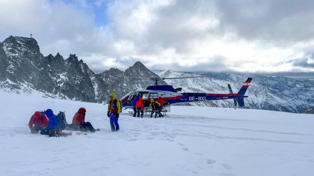 Mehrere Menschen mit Ausrüstung stehen und sitzen im Schnee neben einem Hubschrauber in einer verschneiten Berglandschaft.