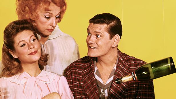 Drei Personen sitzen an einem Tisch, während eine Flasche Sekt in ein rauchendes Glas gegossen wird.