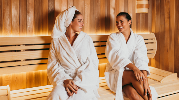 Zwei Frauen sitzen in Bademänteln in einer Sauna und lächeln sich an.
