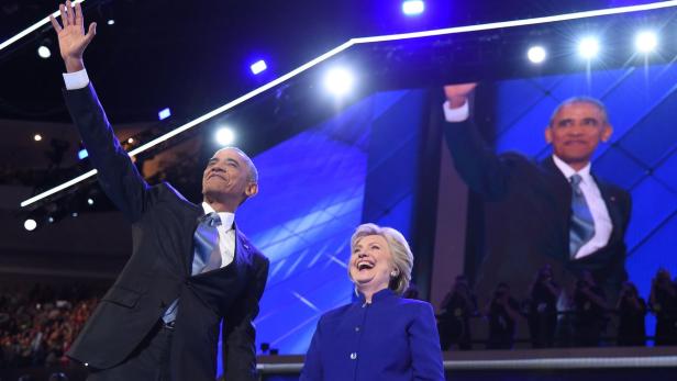 Barack Obama winkt, während Hillary Clinton daneben steht und lacht.