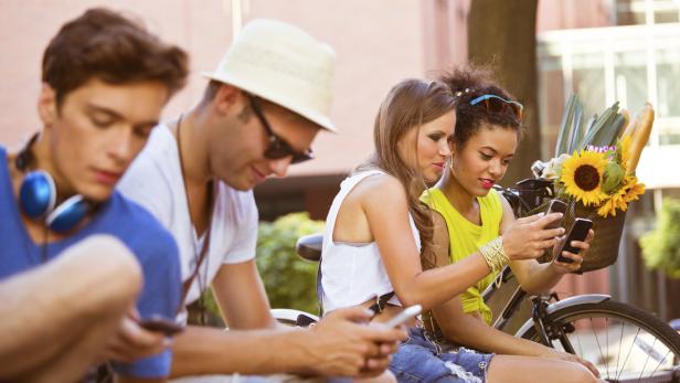 Eine Gruppe junger Leute sitzt im Freien und benutzt ihre Smartphones. Im Hintergrund steht ein Fahrrad mit Blumen und Baguette im Korb.