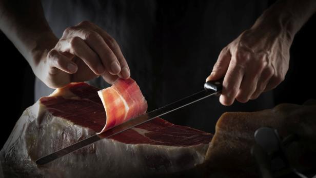 Ein Koch schneidet hauchdünne Scheiben von einem Schinken mit einem Messer.