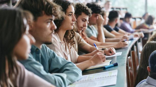 Man sieht junge Studierende in einem Vorlesungssaal. Sie machen Notizen