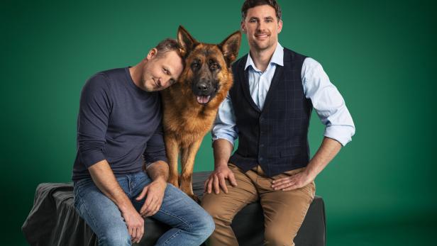 Zwei Männer posieren mit einem Deutschen Schäferhund vor einem grünen Hintergrund.