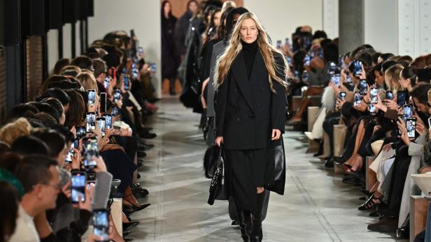 Eine blonde Frau in einem schwarzen Hosenanzug läuft auf einem Laufsteg, umringt von Zuschauern mit Handys.