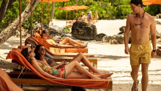Ein Mann geht am Strand entlang, während sich andere unter Sonnenschirmen auf Liegen entspannen.