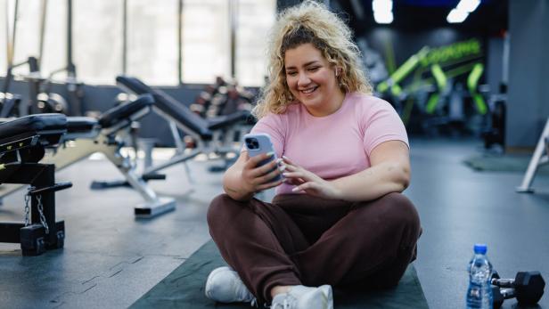 Eine Frau sitzt im Fitnessstudio und schaut in ihr Smartphone.