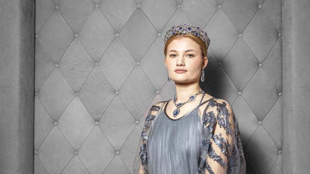 Eine junge Frau sitzt in einem grauen Kleid und Diadem auf einem Sofa.