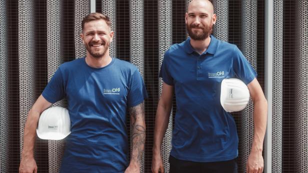 Zwei weiße Männer stehen mit blauen Shirts und weißen Helmen da. 