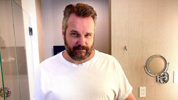 Mann mit Bart steht in einem weißen T-Shirt in einem Badezimmer, die Arme sind von seinem Körper leicht weggestreckt