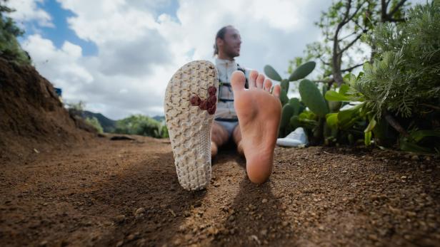 Ein Mann sitzt barfuß auf einem Hügel, ein Fuß und ein Schuh im Vordergrund.
