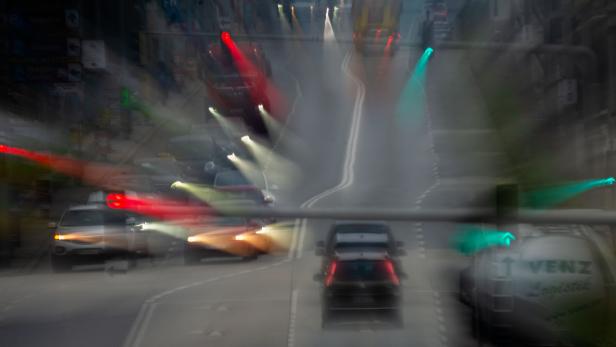 Mehrspurige Straße mit Autos, verschwommenen Scheinwerfern und bunten Ampeln bei Dämmerung oder schlechtem Wetter.