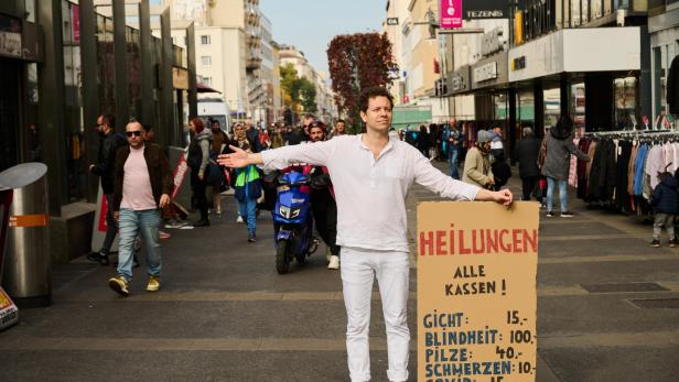 Ein Mann in weißem Gewand in einer Einkaufsstraße. Er hält ein Schild, auf dem er "Heilungen" anbietet.