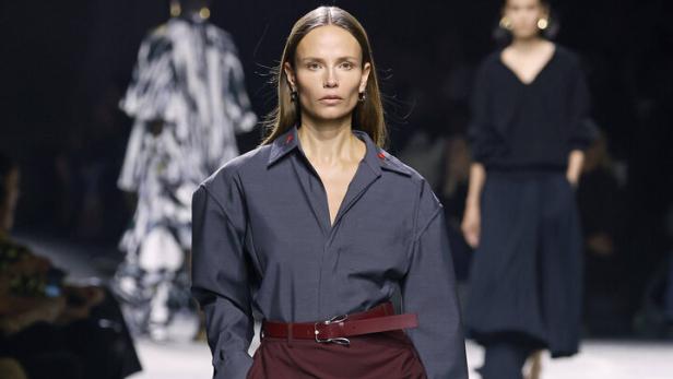 Eine Frau in einem grauen Hemd und einem burgunderroten Rock auf einem Laufsteg mit einer Handtasche.