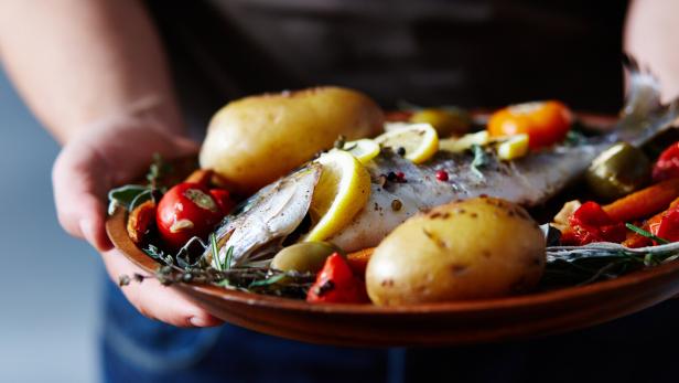 Ein ganzer gebratener Fisch mit Zitronenscheiben, Kartoffeln, Tomaten und Kräutern auf einem Teller.