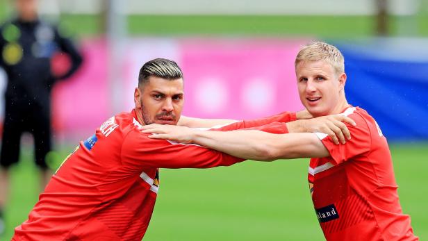Zwei Fußballspieler in roten Trikots dehnen sich gemeinsam auf dem Trainingsplatz.